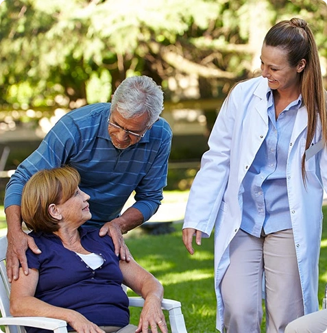 A nurse talks to an elderly woman in a wheelchair with a man beside her outdoors.