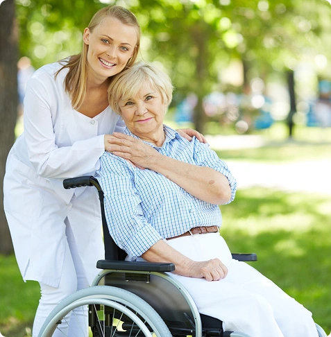 A caregiver embraces an elderly woman in a wheelchair outdoors.